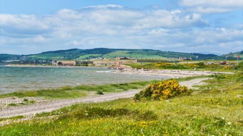 The shoreline of the Inner Moray Firth near Ardersier. A narrow stony path and grass lines the shore. Fort George can be seen in the background.