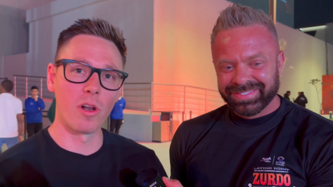 Two men in black T-shirts looking at the camera and talking.