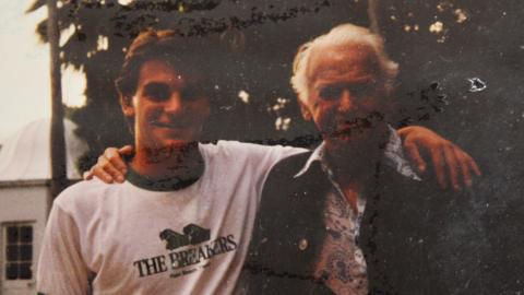 Nick Weston in a t-shirt and Douglas Fairbanks Jr in a floral shirt and jacket posing for a photograph in a garden. They have their arms around each other and are smiling.