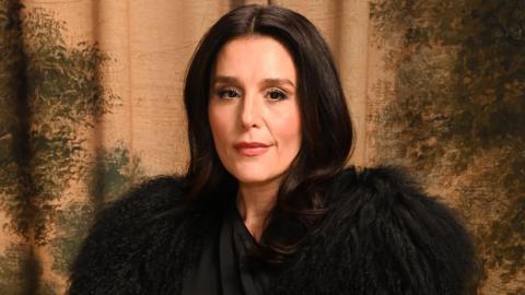 Jessie Ware poses for a photographer. She has a slight smile on her face. She is wearing a dramatic black outfit, covering her shoulders with feathery fabric. She has long, dark hair and is in front of a patterned backdrop of earthy colours.