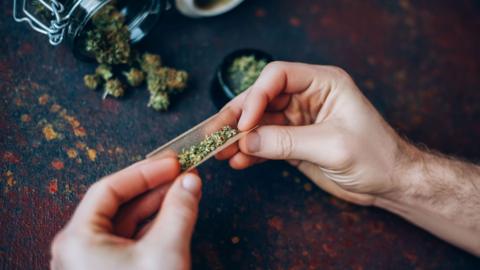 Man's hands twisting marijuana into brown paper. The hands are resting on a dark table and there is a mason jar on its side with marijuana falling out of it.