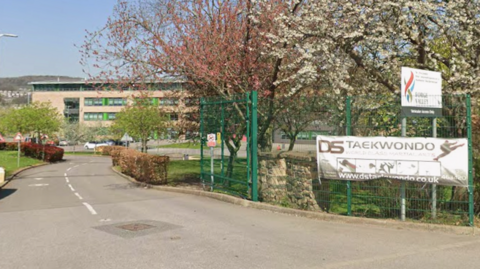 A driveway leads towards a modern building, bordered by trees in blossom, hedges, and green fencing. Signs on the fence include a taekwondo banner and a site sign that appears to read “Forge Valley”. The scene is bright and spring-like, with flowering trees and landscaped grounds surrounding the entrance road.