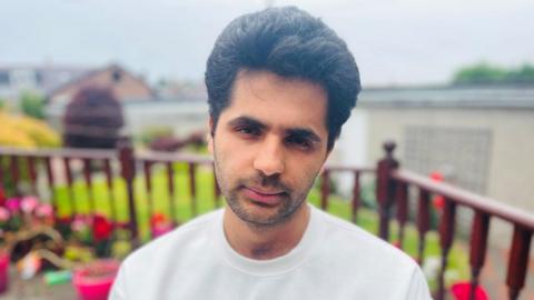 Barun Lamichhane, a man in a white T-shirt, with black hair, looking solemnly at the camera. There is a garden with flowers behind him.