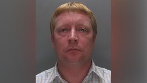 Kevin Perry, with short ginger-blonde hair and wearing a white, blue and green checked shirt, stares at the camera against a grey background