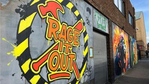 The outside of the venue, which has a graffiti sign on the wall outside with Rage It Out written in orange letters on top of a hammer and baseball bat drawing. A black-and-yellow circle surrounds the writing. To the right is a corrugated metal shutter with the word 'entrance' written in green above it. On the right of the door is another, large bit of street art.