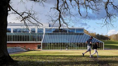 An outside view of the Burrell, taken from the grass shows the glass structure set in a green space and surrounded by woods. Two people, a man and a young girl are jogging past.