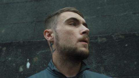 Pete is looking off to his left. He is stood against a grey concrete wall, wearing a dark green fleece. He has short brown hair, a short brown beard and a tattoo of a cross on his neck behind his right ear. 