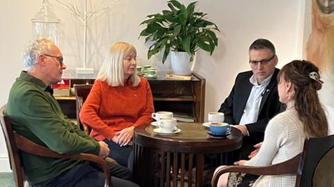 Chris and Callum's parents are sitting around a wooden table in a smart lounge with cups of coffee before them. They are all looking very serious.
