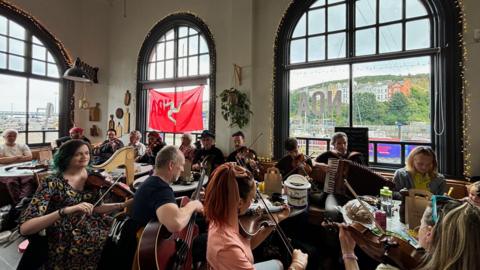 A large group of people sat in a cafe with their instruments including violins, harps and accordian