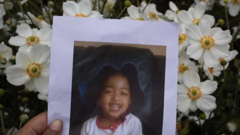 A hand can be seen holding an old photograph of a young girl. She is smiling at the camera. White flowers can be seen behind the photograph.