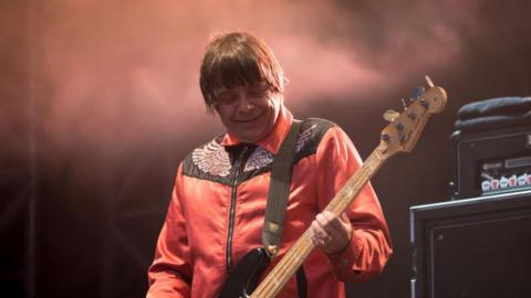 Mani on stage holding his bass guitar on stage. He is looking down at his instrument and smiling - two amps are behind him