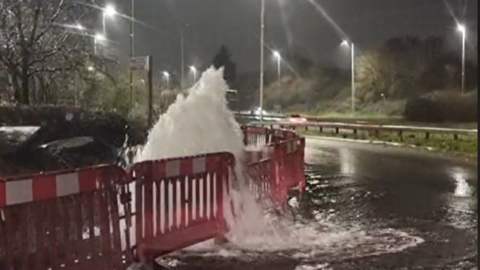 Large amounts of water can be seen coming on to the road with red barricades around it.