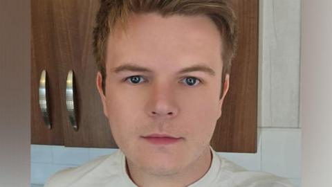 Arron Lewis is smiling. He has short blonde hair and is wearing a white top. He is pictured in what appears to be a kitchen, with kitchen cupboards behind him.