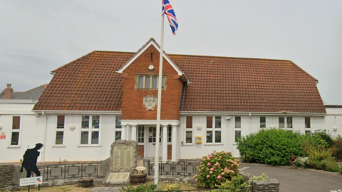 A building with a roof of red tiles and white walls stands among well maintained gardens and with a silhouette of a soldier in front of a war memorial with names on it.