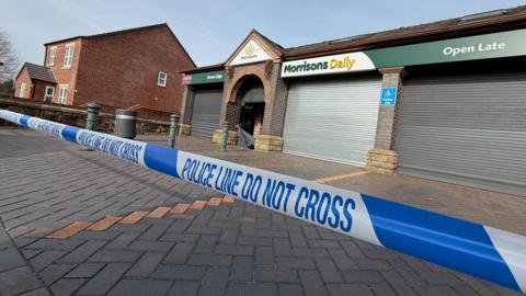 The entrance to a convenience store, with its shutters down. There is police tape over a paved area in front of the store.