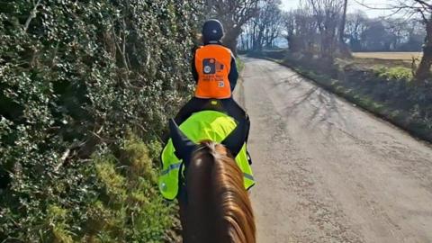 A picture of a horse rider wearing an orange jacket and a black helmet. The image is taken from a horse rider traveling behind.