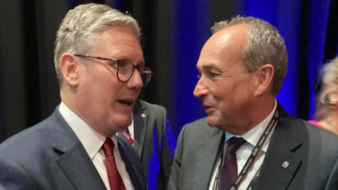 Sir Keir Starmer and chief minister Lyndon Farnham. Sir Kier is wearing a suit, glasses and a red tie.Farnham is wearing a dark grey suit and a grey tie. They are laughing together.