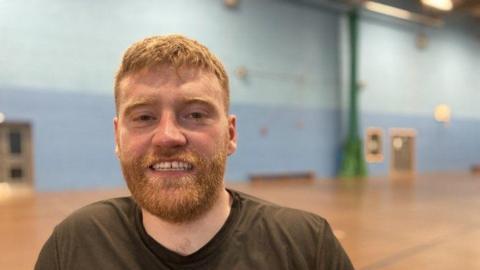 A man with ginger hair and beard is in a sports hall, he is smiling at the camera and wearing a grey t shirt. 