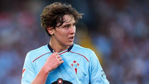 Fer Lopez in action for Celta Vigo. He is wearing a light blue shirt and is holding the neckline with his right hand while looking to his left at the action.