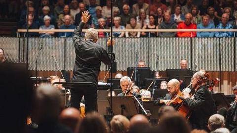 A conductor, dressed in black, raises one hand in the air mid-performance in front of a strings sections. From a raised balcony, an audiences watches the musicians play. 