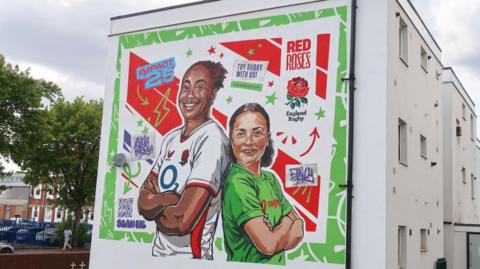 A mural on the side of a building. The wall is white and the painting features a woman in a white England rugby top and a girl in a green rugby top.