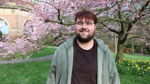 The image shows a young man standing outdoors in a park during springtime. He is smiling at the camera and wearing glasses, a dark t‑shirt, and a green hooded jacket. Behind him, pink blossom-covered trees are in full bloom, with green grass and yellow flowers visible at ground level.