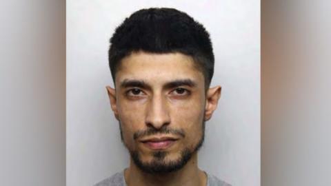 Yasar Hussain: A dark-haired man with a dark beard wearing a grey round-necked t-shirt. He is appearing in his police custody photo.