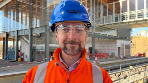 Stephen Deaville is standing on the station construction site wearing a blue hard hat, safety glasses and an orange high visibility coat. Its is a sunny day with blue skies. He has blue eyes and facial hair. Behind him is the incomplete station.