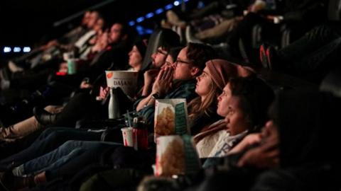 People in a row in a multiplex cinema. They are looking at the screen with popcorn and soft drinks