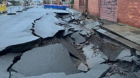 Grey temporary fencing and fluorescent barriers surround the sinkhole, pictured in January. The grey tarmac road is splintered and sunken.
