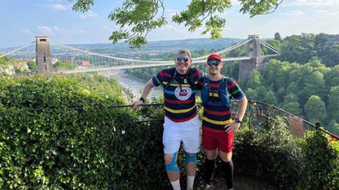 Simon Pilkington, pictured right, stands next to Rob Dowling, pictured left, in front of the Clifton Suspension Bridge. They are both wearing tops with black, red, yellow, blue and green stripes. 
