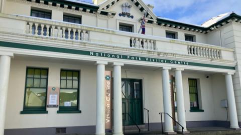 Exterior of the national park visitor centre. The building is white with green paint. Pillars are in front of the green entrance.