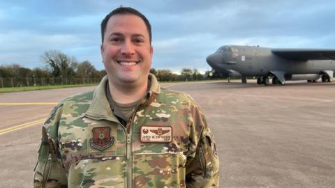 Lieutenant Colonel Jared Patterson wearing his US Air Force camouflage uniform, smiling at the camera as a B-52 bomber stands on the runway behind him