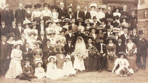 A sepia picture of a a wedding party outside a place of worship. The bride can be seen sitting in the centre of the photograph, wearing a white dress and a veil. Attendees are wearing hats and holding flower bouquets.