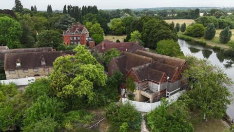 A drone view of the hotel site, fenced off and surrounded by large trees. The River Thames is to the right of the buildings with fields in the background