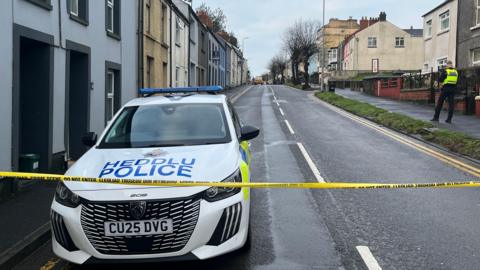 A police car parked on the road. In front is yellow police tape. To the left and right are a row of terraced houses. There is a police officer in hi-vis walking on the right of the street.