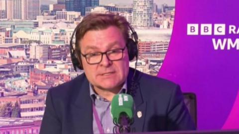 John Cotton is sitting on a chair in front of a green microphone with headphones on. He is sat against a BBC Radio WM purple backdrop with the skyline of Birmingham in the background.