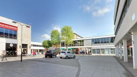 Modern shopping square with two-story concrete building with glass frontages, with pillars in front of the buildings to the right. There are two cars, black and white, parked in the grey-paved square. A few people are seen walking through the square.