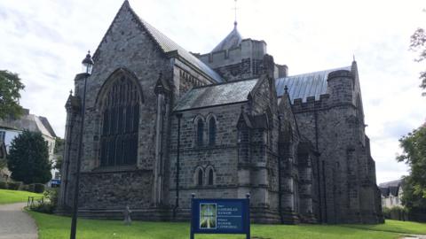 A picture of the front of the large Bangor Cathedral on a bright day with a blue sign at the forefront of the picture