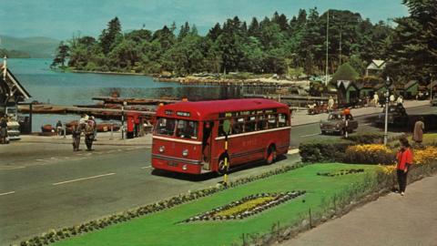 Postcard dating from the 1960s showing - in highly saturated colour - a red single decker bus on a road next to the shore of a lake. A number of pedestrians are strolling on the pavement, next to a floral display in a grassed area and there are rowing boats tied to jetties on the lake.