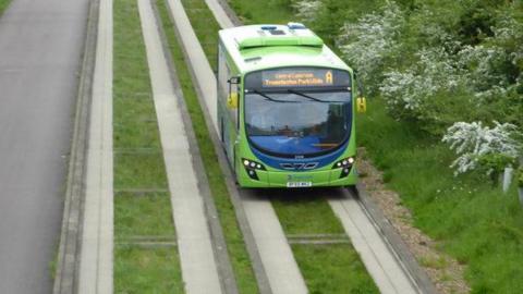 A Cambridge Busway vehicle heading towards us on the right hand side of the Busway, with a verge and shrubbery on the right and a normal road on the left.