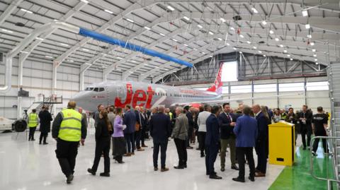 Dozens of guests standing in the new hanger. They are gathered next to a Jet2 plane, which has a grey and red livery and the firm's logo on the side.