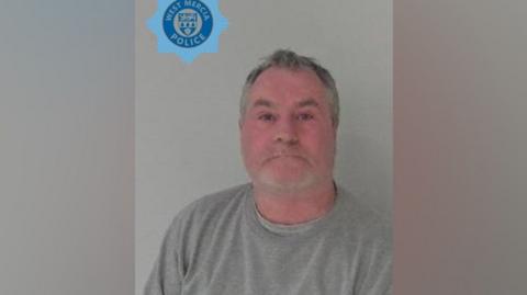 Dave Butler in a police photo. He is wearing a grey top and looking at the camera. He has grey hair and grey stubble and the West Mercia Police logo is printed on the photo to the top left. 