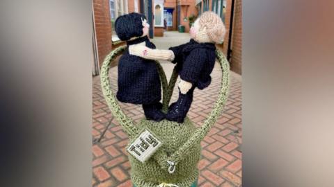 Two woollen figurines hold hands with a green knitted heart around them, atop a bollard in a pedestrianised shopping street