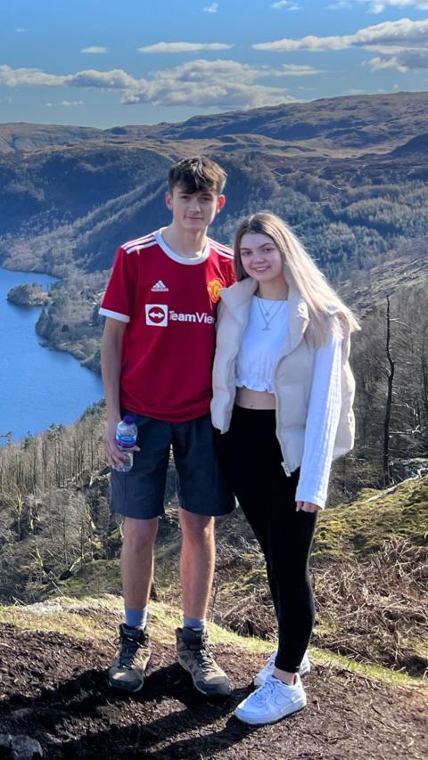 Alt text: A scenic hillside photo showing **Lloyd** on the left, wearing a red Manchester United shirt, dark shorts and hiking boots, holding a water bottle, and **Louise** on the right, wearing a white long‑sleeve top, beige vest, black trousers and white trainers. They’re standing side by side overlooking a lake and rolling green hills under a bright, partly cloudy sky.