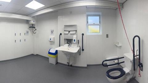 The large white bathroom, which has a toilet with handles, a red cord for assistance, a hoist is in the corner, and handles around the sink.