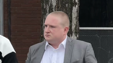 A man with a shaven head wearing a grey blazer over a white shirt standing in front of a brick wall