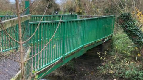 A footbridge with a green railing. It is leading over a brook that is overgrown with trees and bushes.
