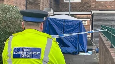 A police community support officer stands in front of a blue forensic tent.
