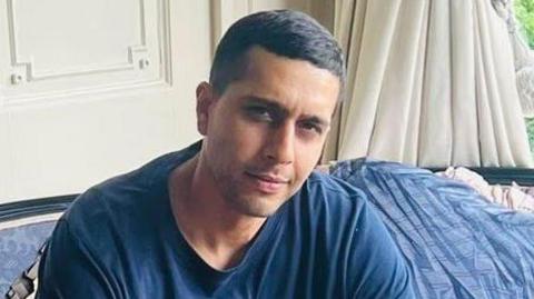 Kamran Rasool Aman, looking at the camera. His head is slightly tilted to one side, and he wears a blue t-shirt and has short black hair.